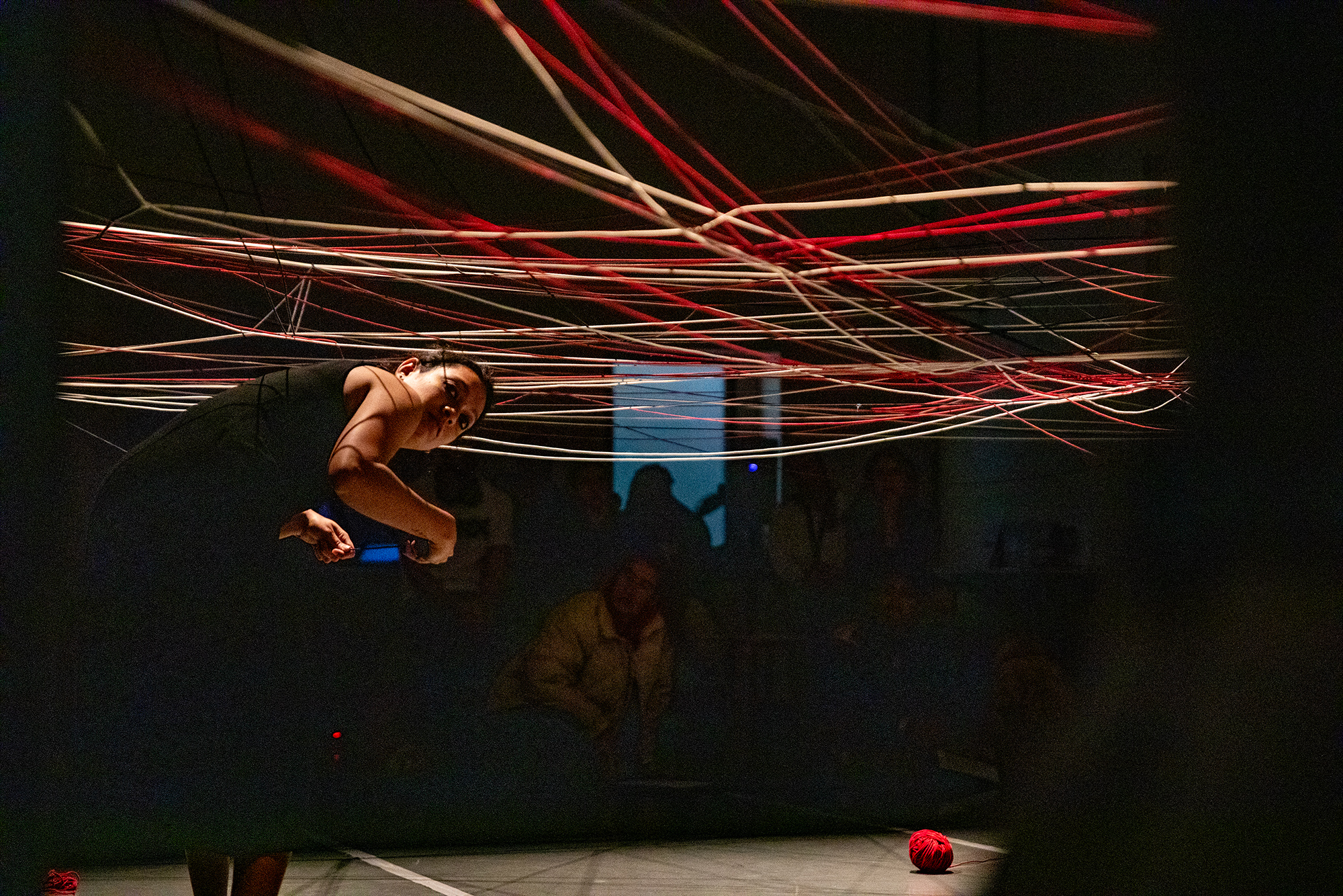 Priyakshi Agarwal ist standing bent over on stage. Above her is a net made of stretched yarns. A ball auf yarn lies in front of her. The spot light highlights the stage leaving the are beside in the dark.