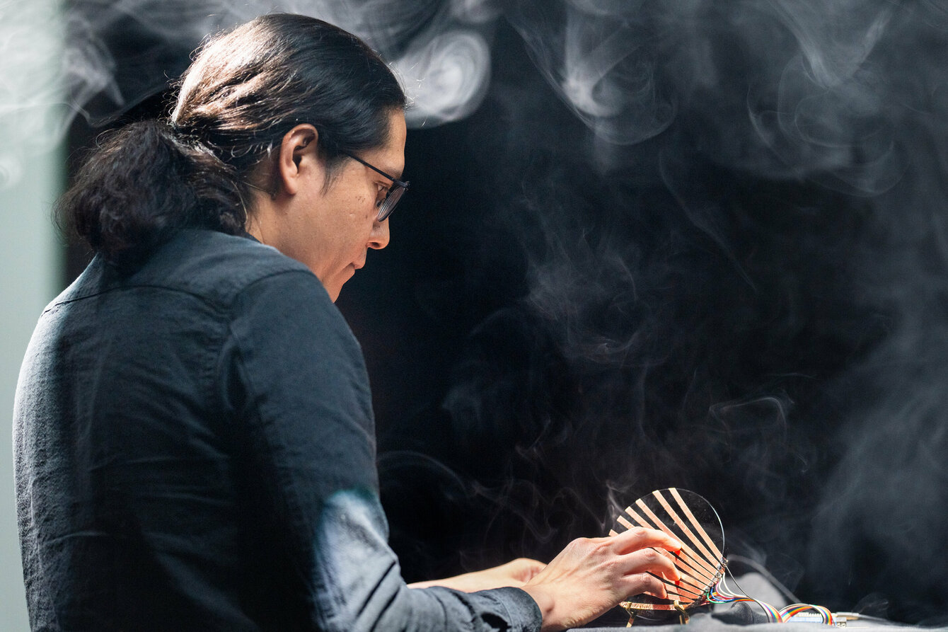 Eine Person berührt einen kunstvoll gestalteten elektronischen Sensor, der auf einem hohen Tisch liegt. Auf der Bühne, die beleuchtet ist, liegt Nebel in der Luft. 