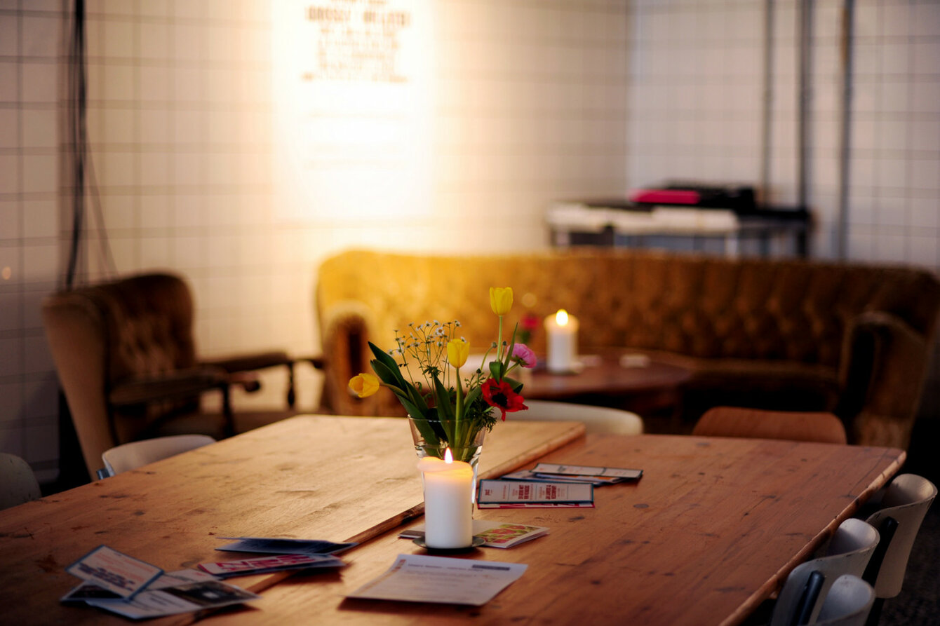  Das Bild zeigt einen gemütlichen Innenraum des Foyers von PACT Zollverein. Im Vordergrund steht ein Holztisch, auf dem eine brennende weiße Kerze in einem Glasgefäß und ein kleiner Blumenstrauß mit gelben Tulpen und roten Blumen arrangiert sind. Rund um den Tisch liegen einige Prospekte oder Flyer verteilt. Im Hintergrund sind ein beiger, gepolsterter Sofa und ein brauner Sessel zu sehen. Die Beleuchtung ist warm und schafft eine einladende Atmosphäre. 