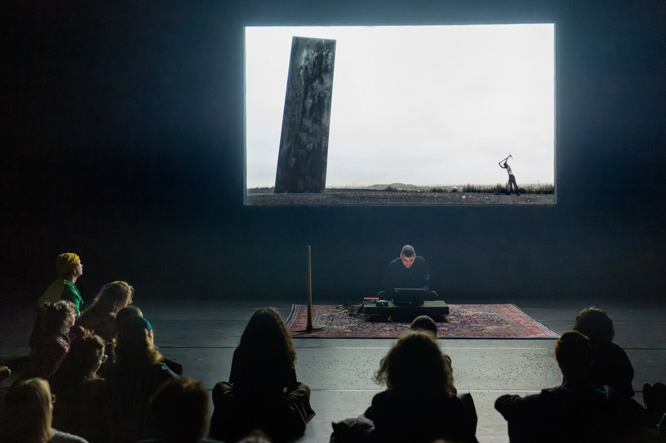 A performer sits on the floor on a carpet and uses a laptop to control the video projection behind him. The audience sits in a semi-circle around him.