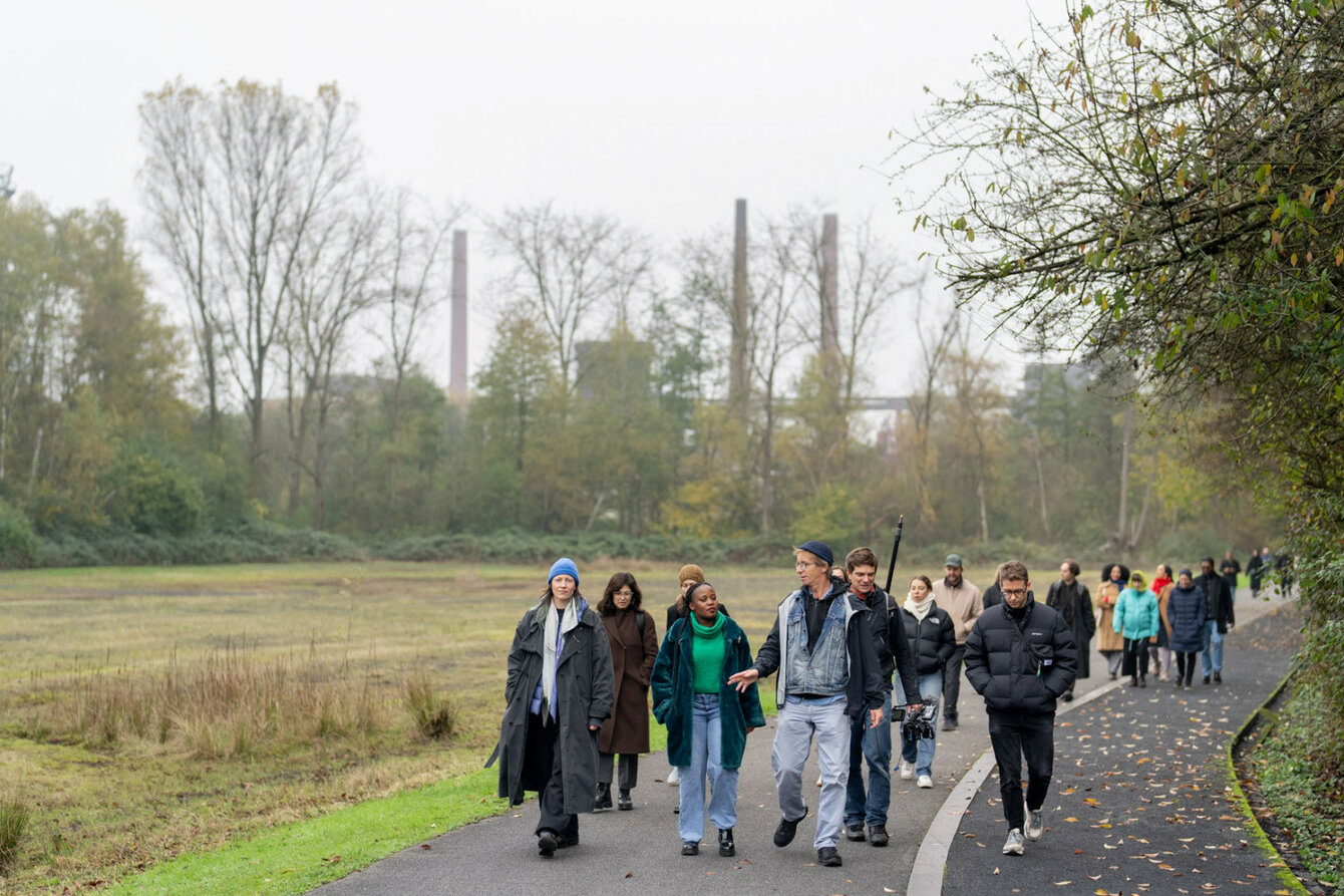 A group of people are walking along a green path. The group walks towards the camera.