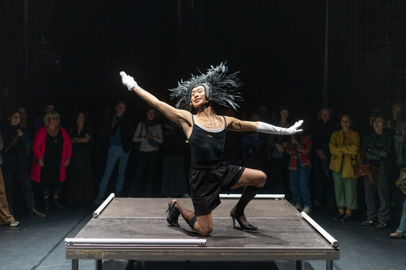 A person in a feather wig strikes a dancing pose on a small stage and smiles. The audience is all around and in the dark.