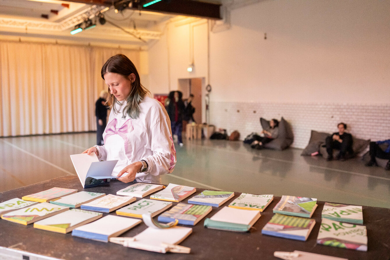 A woman is reading in the foreground. She is standing at a table reading a book with more self-bound books on it.