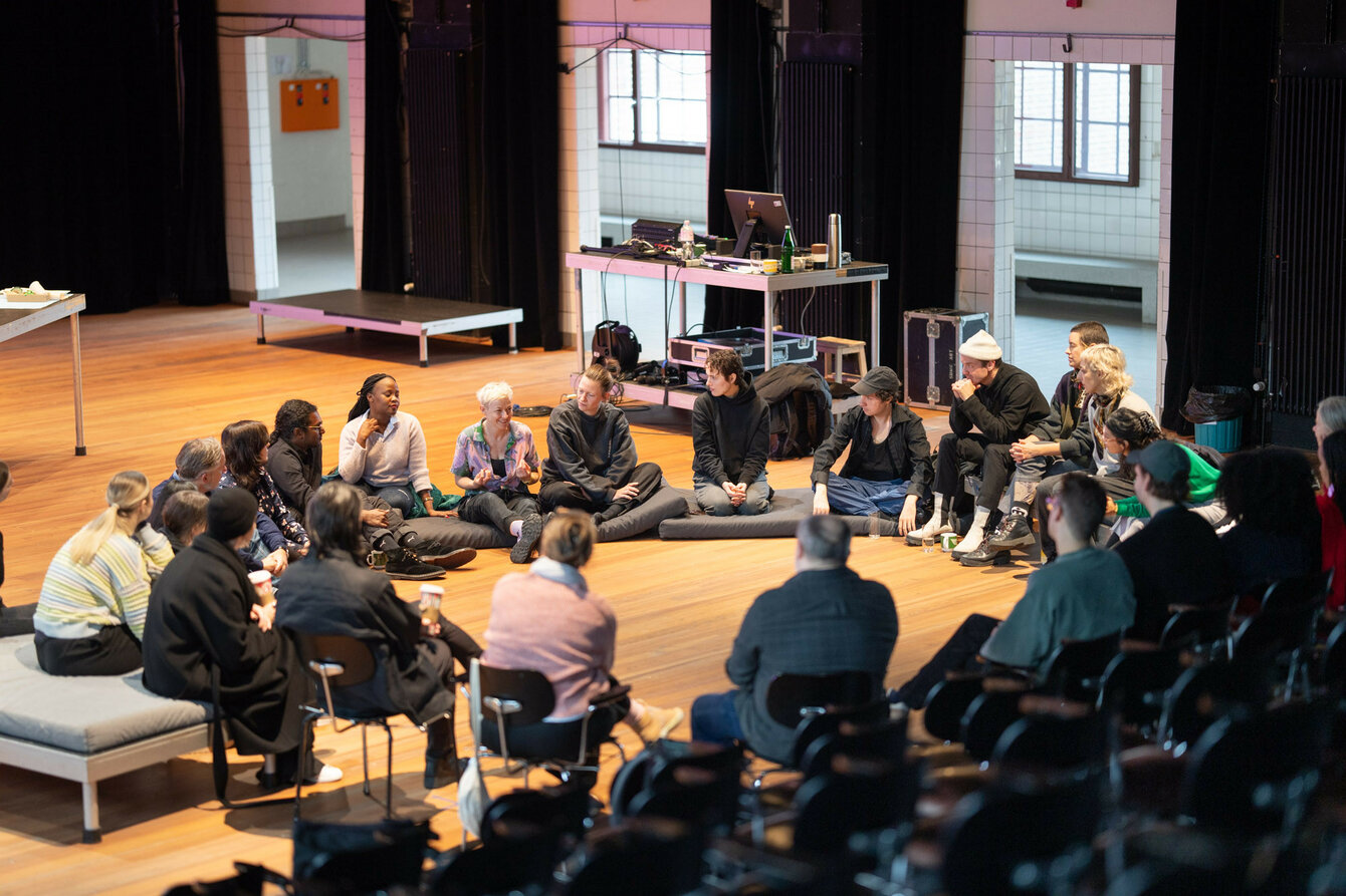 A group of people sit together in a circle and talk. They are sitting in the wooden ground of theatre stage.