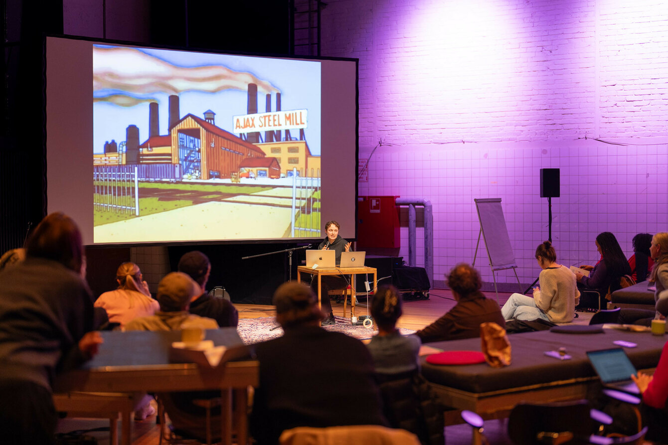 A person gives a lecture on a stage. In the foreground are spectators. A comic-style image of a factory is projected in the background.