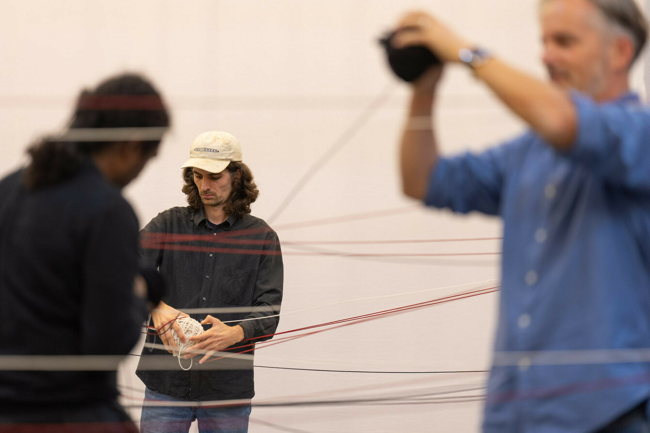 Three people stand between taut threads of yarn in front of a plain background