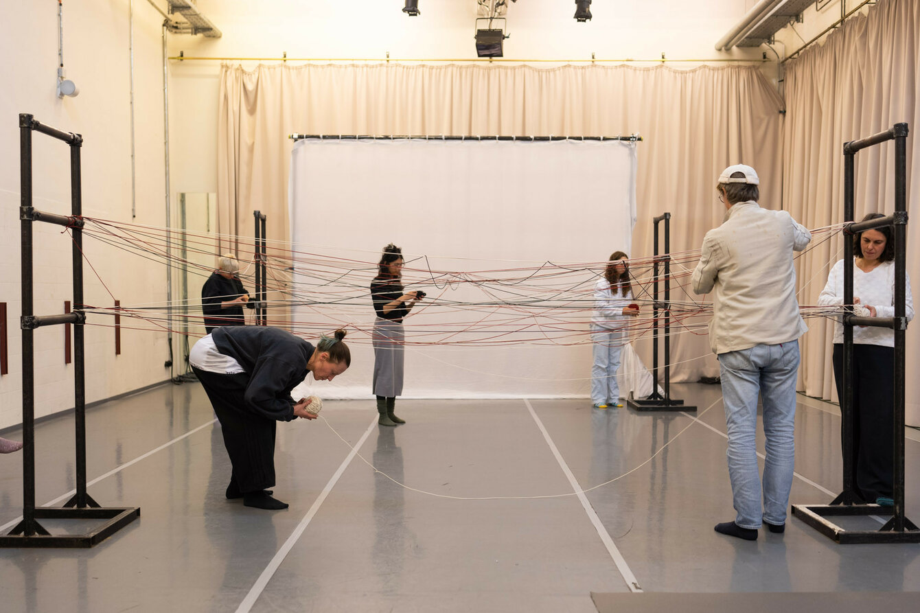 In the dance studio there is an installation made of metal pipes and strings stretched between them. Several people stand next to it and interact with the structure. 