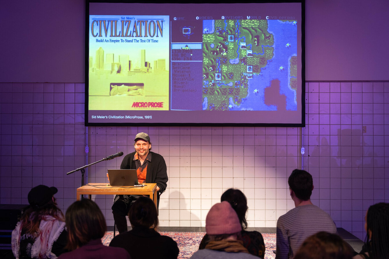 Sebastian Quack gives a lecture on a stage. Computer games can be seen on a projection in the background.