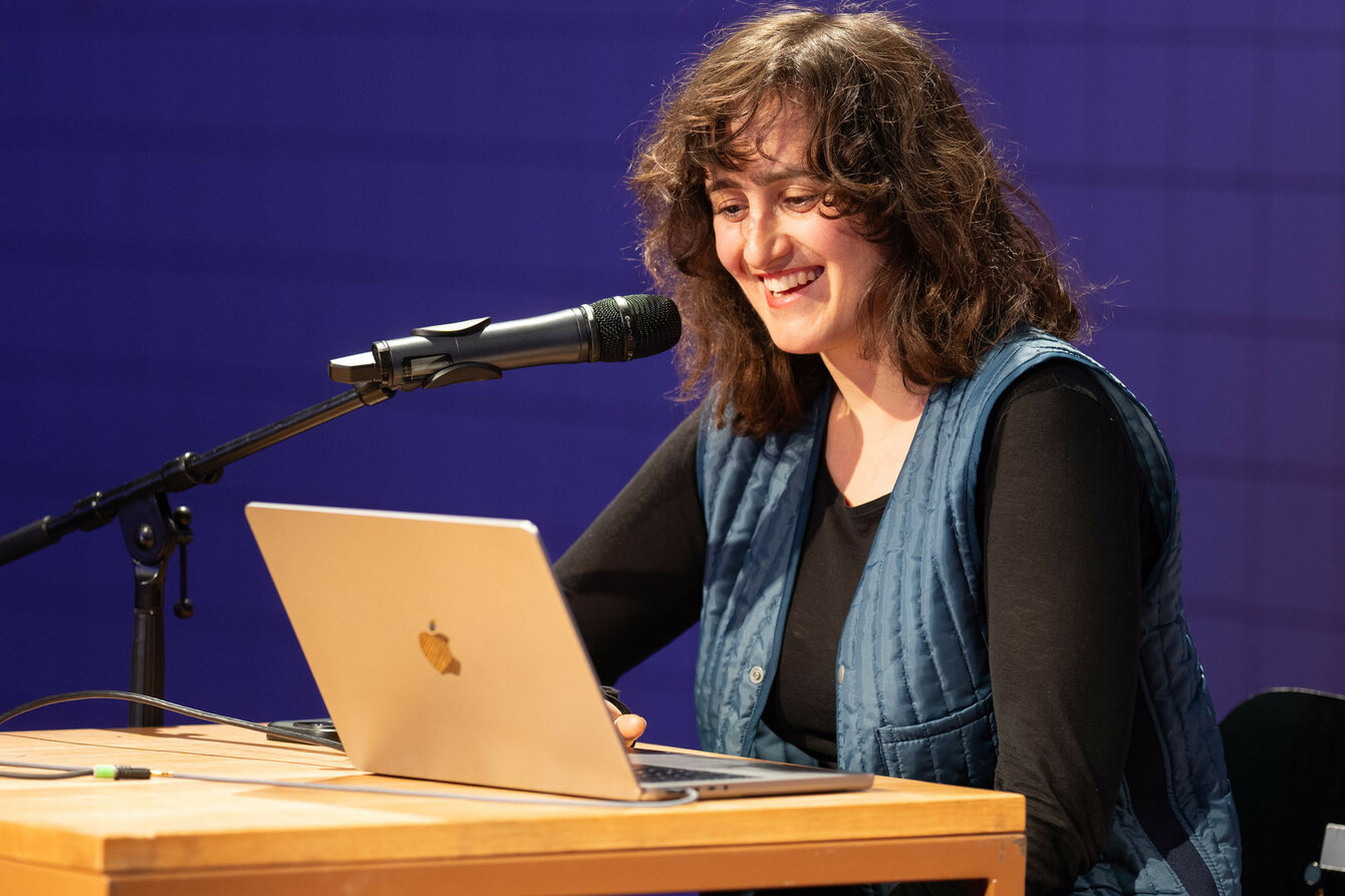 Maria Lisogorskaya gives a lecture on Assemble projects. She is sitting at a table with a microphone and a laptop in front of her.