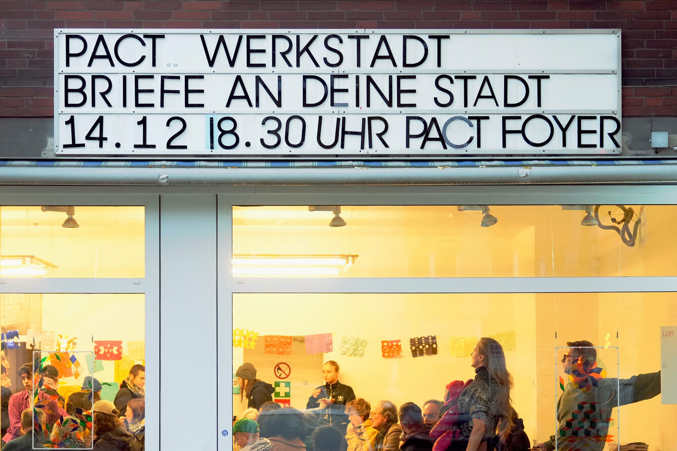 Das Bild zeigt die Außenansicht eines Veranstaltungsortes mit großen Glasfenstern. Oben an der Fassade ist ein Schild mit der Aufschrift „PACT WERKSTADT BRIEFE AN DEINE STADT 14.12 | 18.30 UHR PACT FOYER“ angebracht. Durch die Fenster ist eine Veranstaltung zu sehen, bei der Menschen an Tischen sitzen, sich unterhalten und anscheinend gemeinsam arbeiten oder basteln. Der Raum ist hell beleuchtet, mit bunten Dekorationen an den Wänden, darunter farbige Papiermotive.