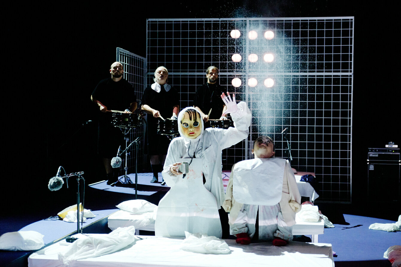 Eine Szene aus NÔT: Zwei Performer*innen, die zwischen zwei Betten auf einer dunklen Bühne sitzen. Der*die Performer*in links ist maskiert und in weiß gekleidet. In der einen Hand ist eine Tasse zu sehen, die linke Hand in der Luft. Daneben eine kleine Person, die mit nach hinten geworfenem Kopf Wasser in die Luft pustet. Im Hintergrund drei schwarz gekleidete Musiker*innen, die vor einer Gitterwand stehen, an der Scheinwerfer zu sehen sind.