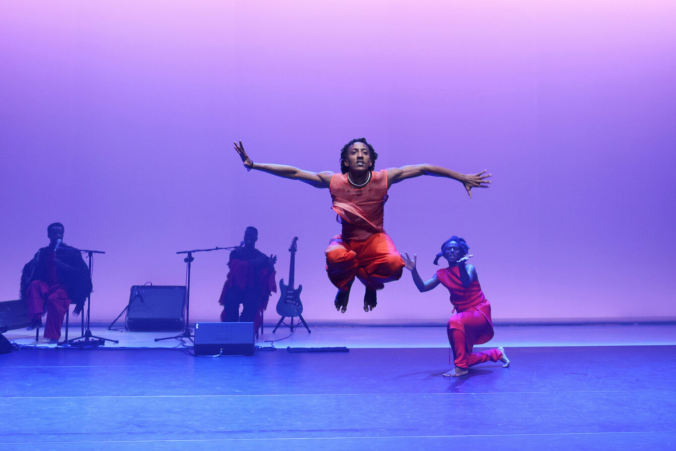 Purple light on the stage. In the middle, a performer jumps into the air with outstretched arms. People sit in the background and watch. One of them points to the person jumping.