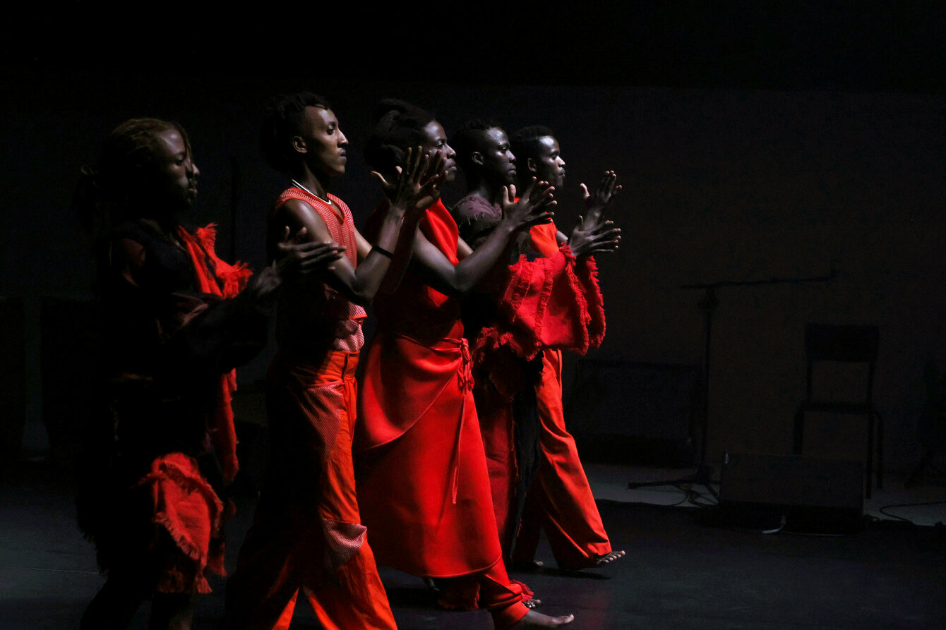 A dark stage, several black performer walking side by side. They are wearing red clothes. They beat their hands together in front of their chests.