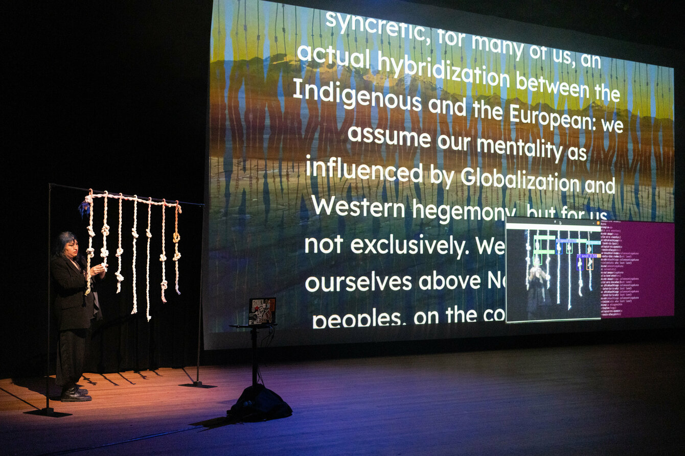 A performer on a dark stage. Next to her is a large screen onto which a text manifesto is projected. The performer next to her is tying khipu knots.  