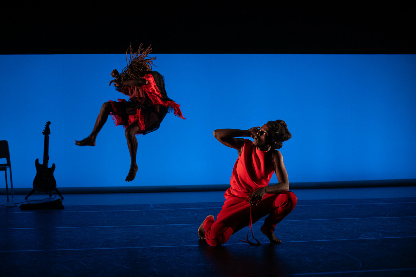 A dark stage with blue backlighting. Two black performers are on stage. They are wearing red clothes. One kneels in the foreground and looks to the side with one hand by his ear. The second one jumps into the air behind. 