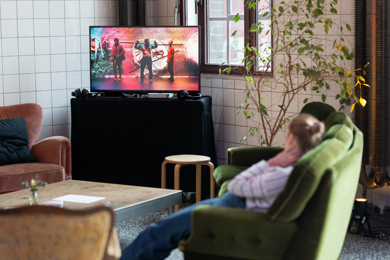Two people sit on a couch and watch a video on a television in a tiled foyer.