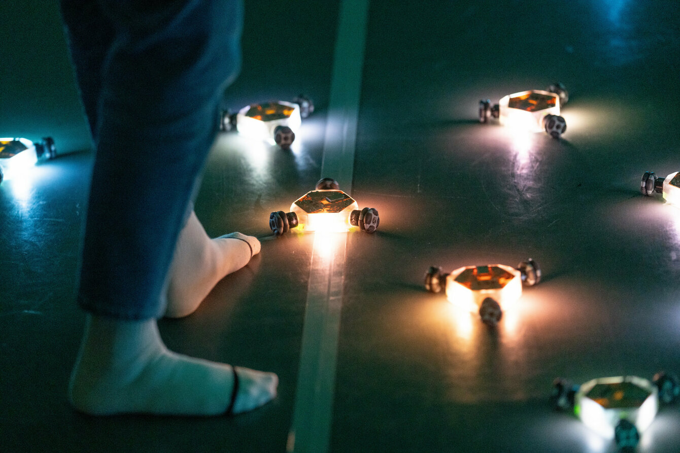Three-wheeled, tennis ball-sized, colorful, flashing robots drive in a joint formation around a person moving past them in socks. 