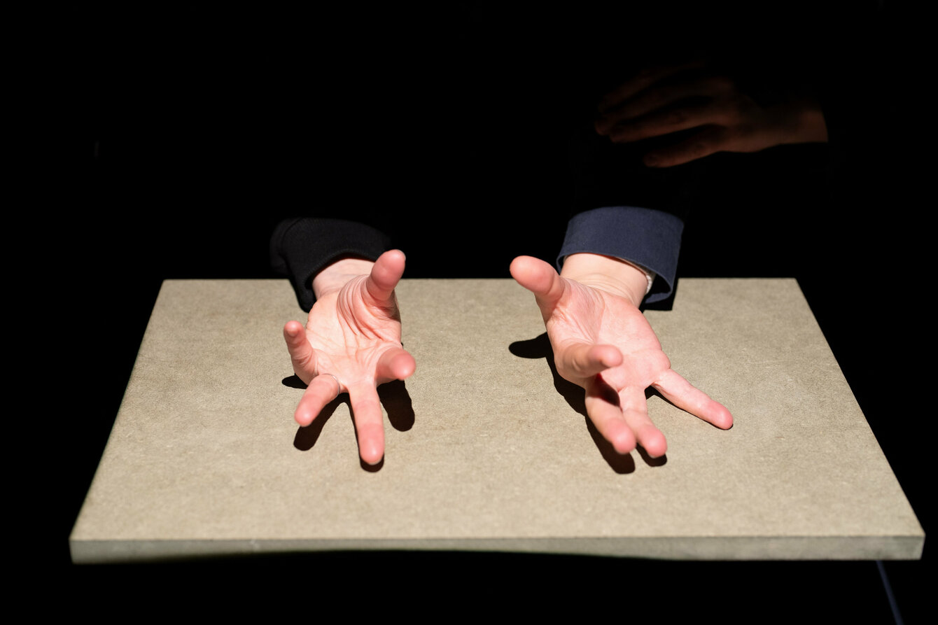Zwei Hände liegen auf einer Spanplatte auf, die Handflächen zeigen nach oben, die Finger sind auseinander gestreckt. Hartes Licht von oben lässt sie scharfkantige Schatten auf den Untergrund werfen. 