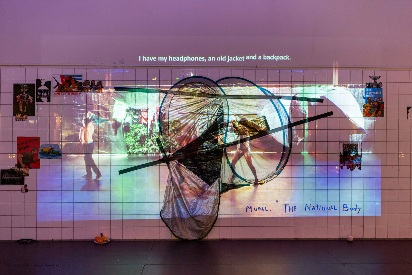 An art installation that is attached to the wall. It consists of a tent which is stuck on with gaffer tape and several photographs stuck around it. A video is projected above and overlapping them.