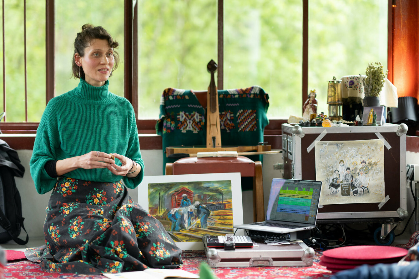 A performer squats in front of objects that she will soon be using in her artistic work: A painting, a pontic lyre and a laptop with music software. 