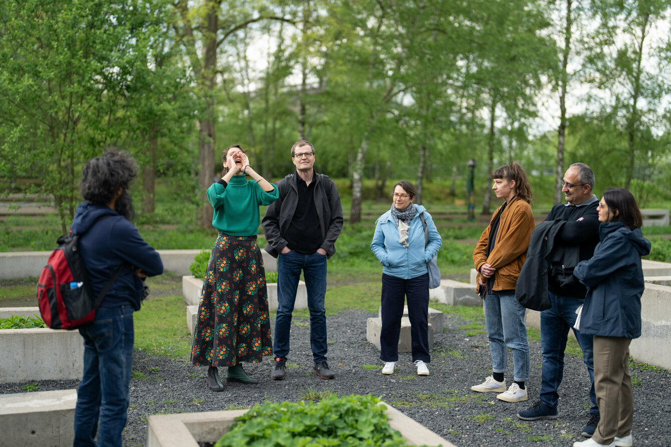 A group of people are standing outside in a herb garden. The performer has placed her hands in front of her mouth to amplify the sound and is singing fervently.