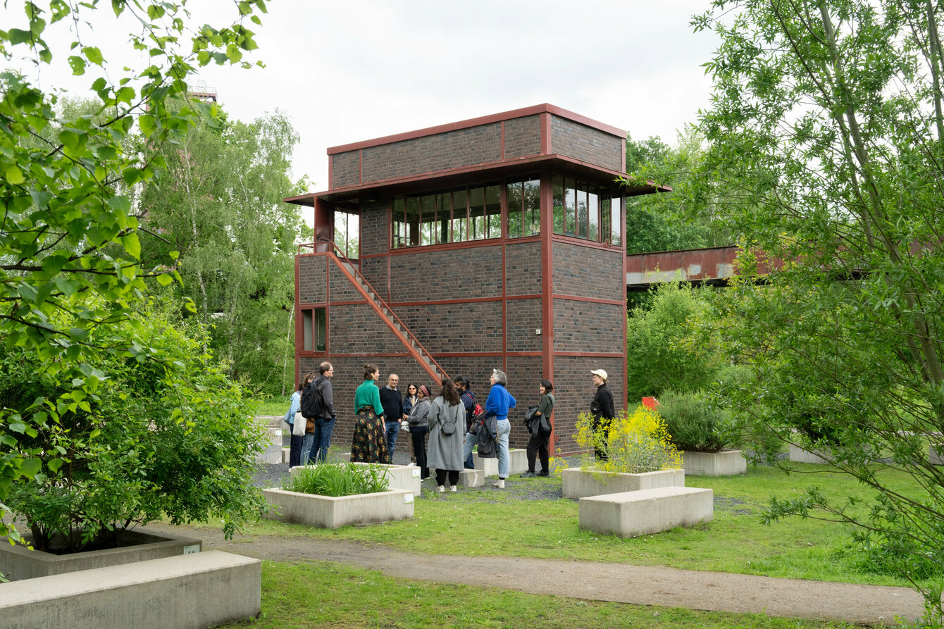In the center of the picture, a group of people are standing in front of the old signal box building at Zollverein, framed by bushes and trees at the edge of the picture. 