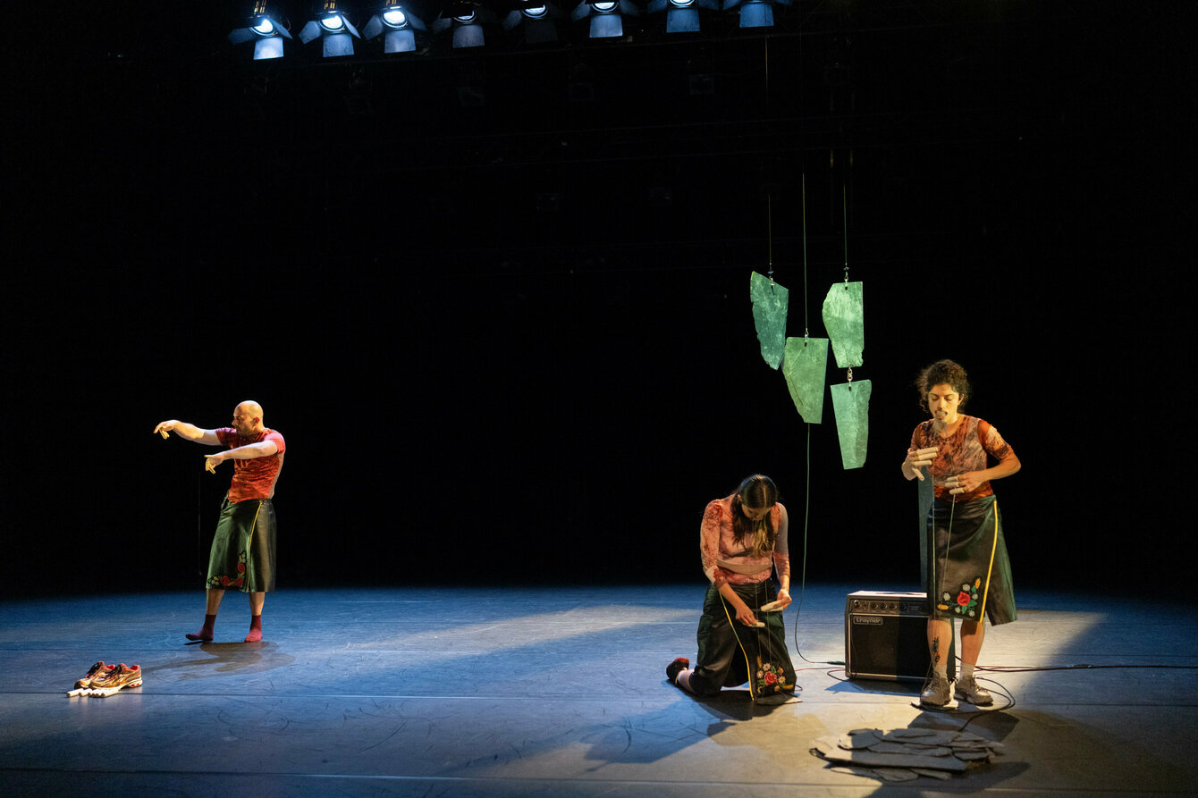Drei Performer:innen auf der Bühne in blauem und grünem Licht. Vorne spielen zwei von ihnen Musik auf elektrisch verstärkten Metallsaiten. Im Hintergrund bewegt sich dazu die dritte Person.