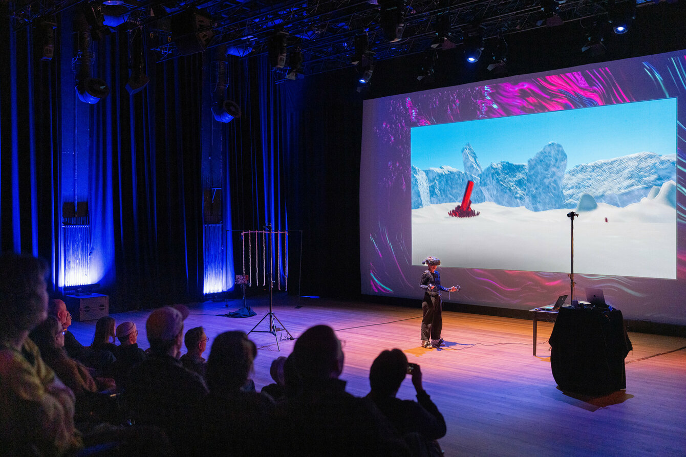 Eine Person steht mit einer VR-Brille auf einer Bühne vor einem Publikum. Hinter ihr wird die virtuelle Welt, in der sie sich befindet, auf eine Leinwand projiziert.