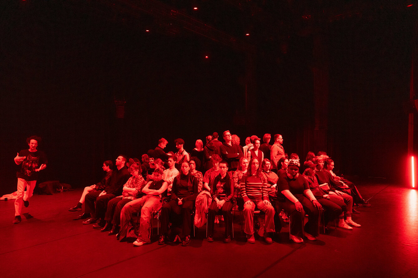 A crowd has gathered in a circle of chairs facing outwards and is looking outwards. A performer moves around them in a circle. It is dark and illuminated in red.