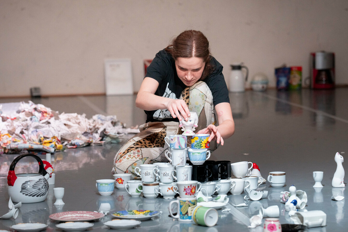 A performer squats on the floor and carefully stacks many individual porcelain cups and plates on top of each other.
