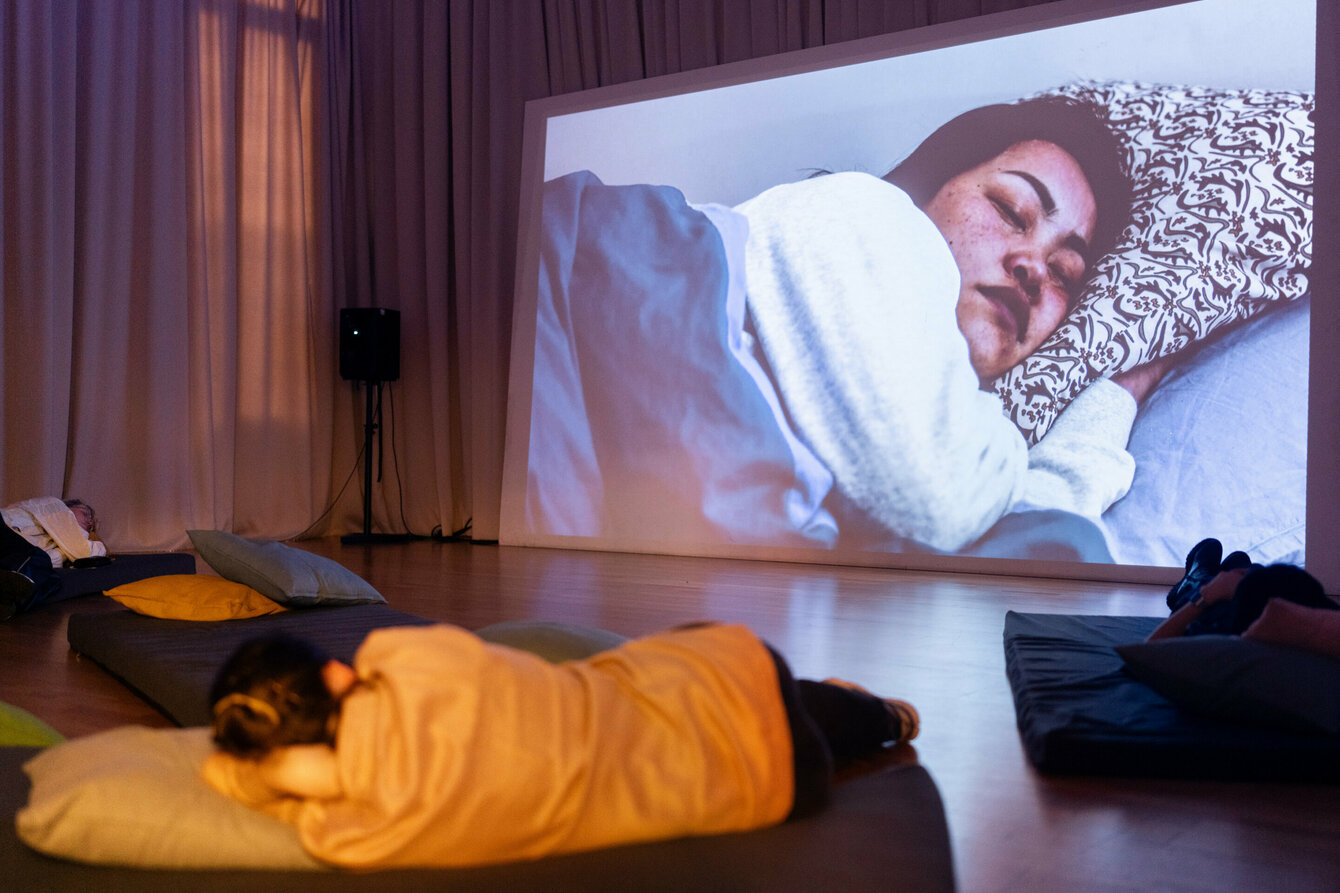 A large film projection is projected onto the wall, showing a person sleeping with their head on a pillow. The viewers of the film are also lying in a similar position in the room on pillows and comfortable underlays.