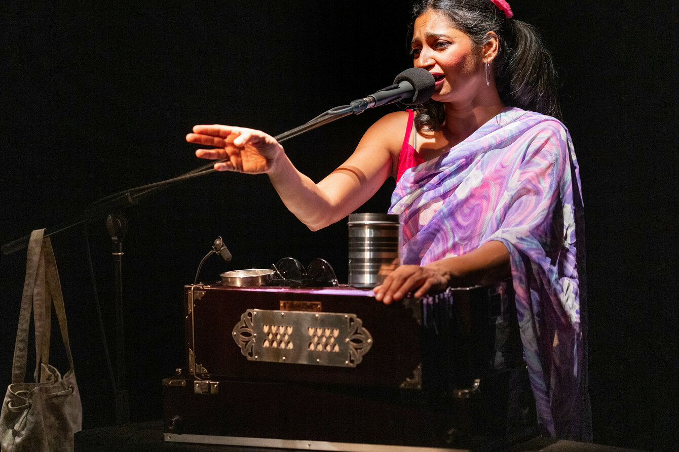 A woman on a stage at the microphone. She wears a purple robe and plays an Indian harmonium.