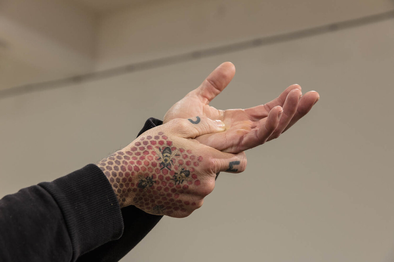 Two hands on a plain background. The thumb of the right hand presses into the open palm of the left hand. The back of the hand is tattooed with a pattern.