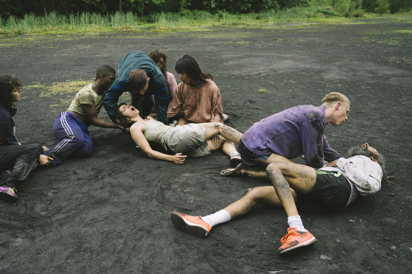 Gruppe von Tänzer:innen liegen mit geschlossenen Augen und geöffneten Mündern auf grauem Schotterboden einer Halde dem Boden im Wald