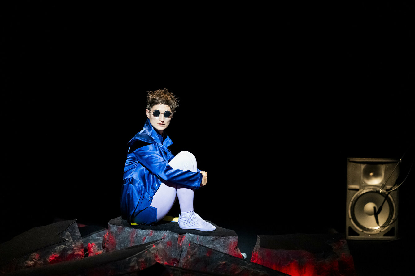 Bühnenbild aus Lava eine Person mit Sonnenbrille sitzt mit angewinkelten Beinen auf einem Stein