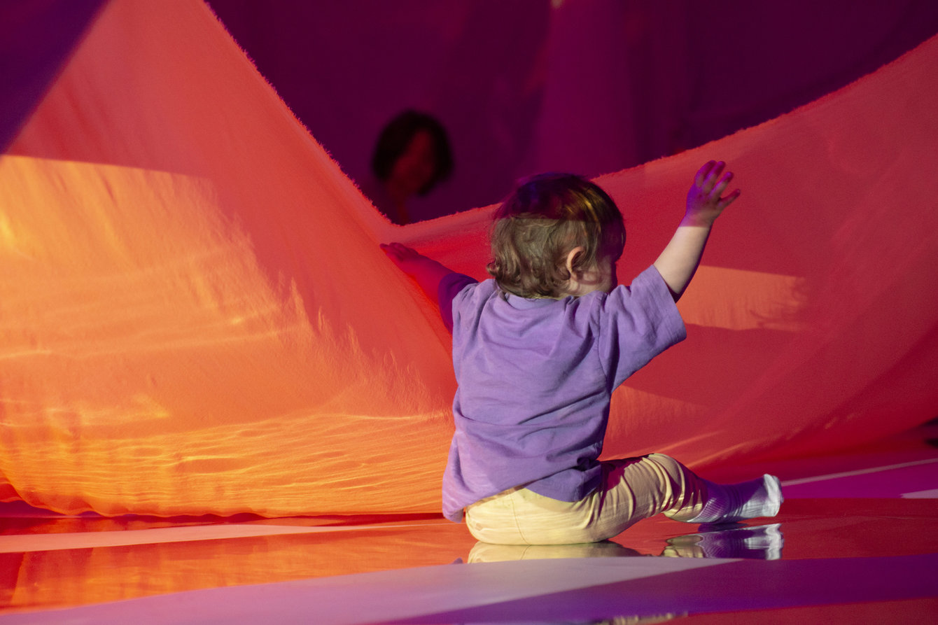 Ein Kind sitzt auf dem Boden und streckt die Hände hoch, ein großen Tuch aus Ballonstoff im Hintergrund