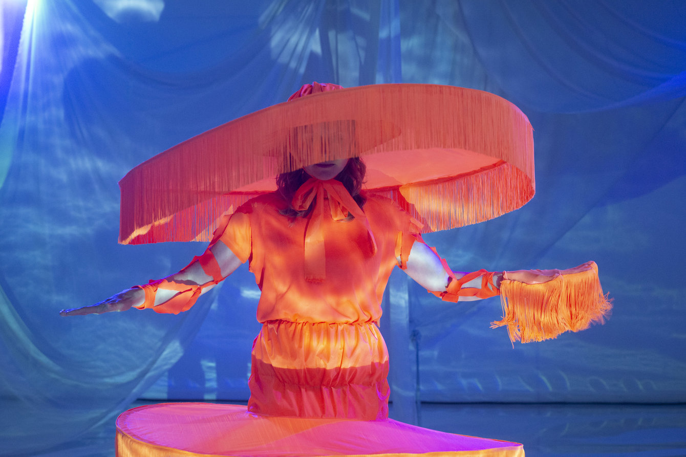 Eine Figur mit Kostüm in Orange und rieseigem runden Hut, der an einen Lampenschirm erinnert vor blauem Hintergrund.