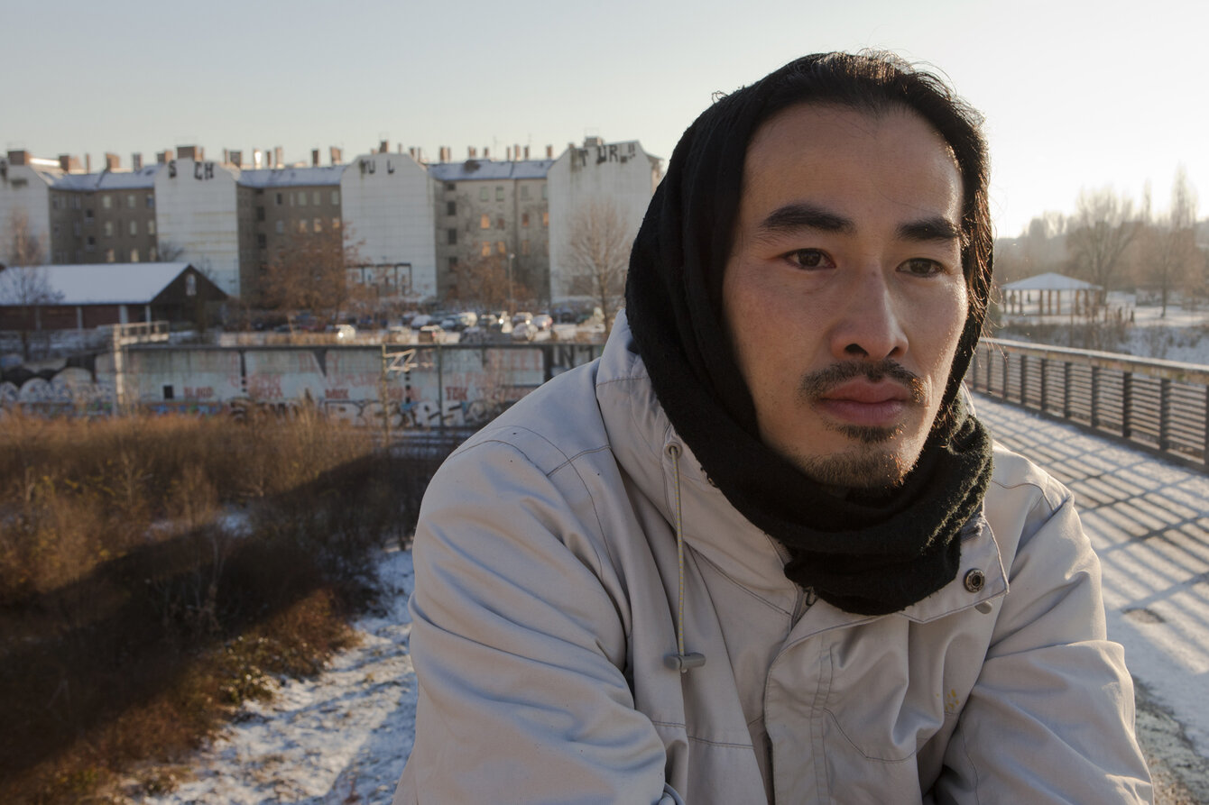 Portrait, der Mensch trägt eine helle Jacke, den Kopf umhüllt eine dunkle Kapuze. Auf die linke Gesichtshälfte fällt fahler, winterlicher Sonnenschein. Im Hintergrund ist eine Wohnsiedlung zu sehen.