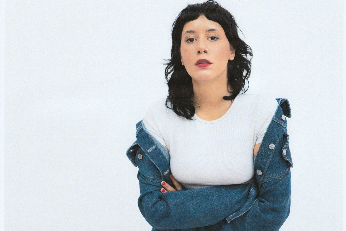 Musician Güner Künier poses in front of a white background and looks into the camera. She is wearing a white top and an open denim jacket over her shoulders.