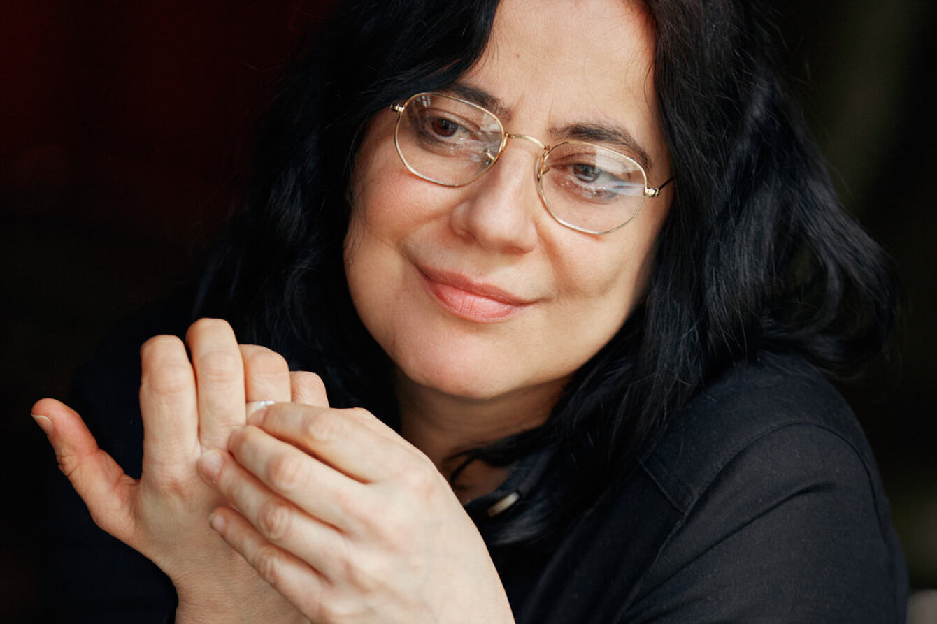 PPortrait of Lütfiye Güzel against a dark background. She is wearing a black top and has her arms resting on a table. Her hands are touching in front of her face. She is smiling.