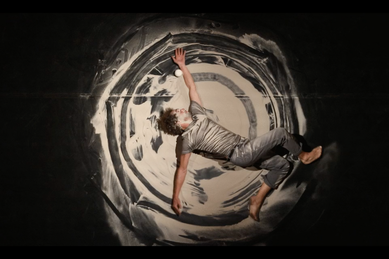 Photography from a bird's eye view. Juggler Tobias Piero Dohm lies on the ground in the sand. Traces of his round movements can be seen in the sand.