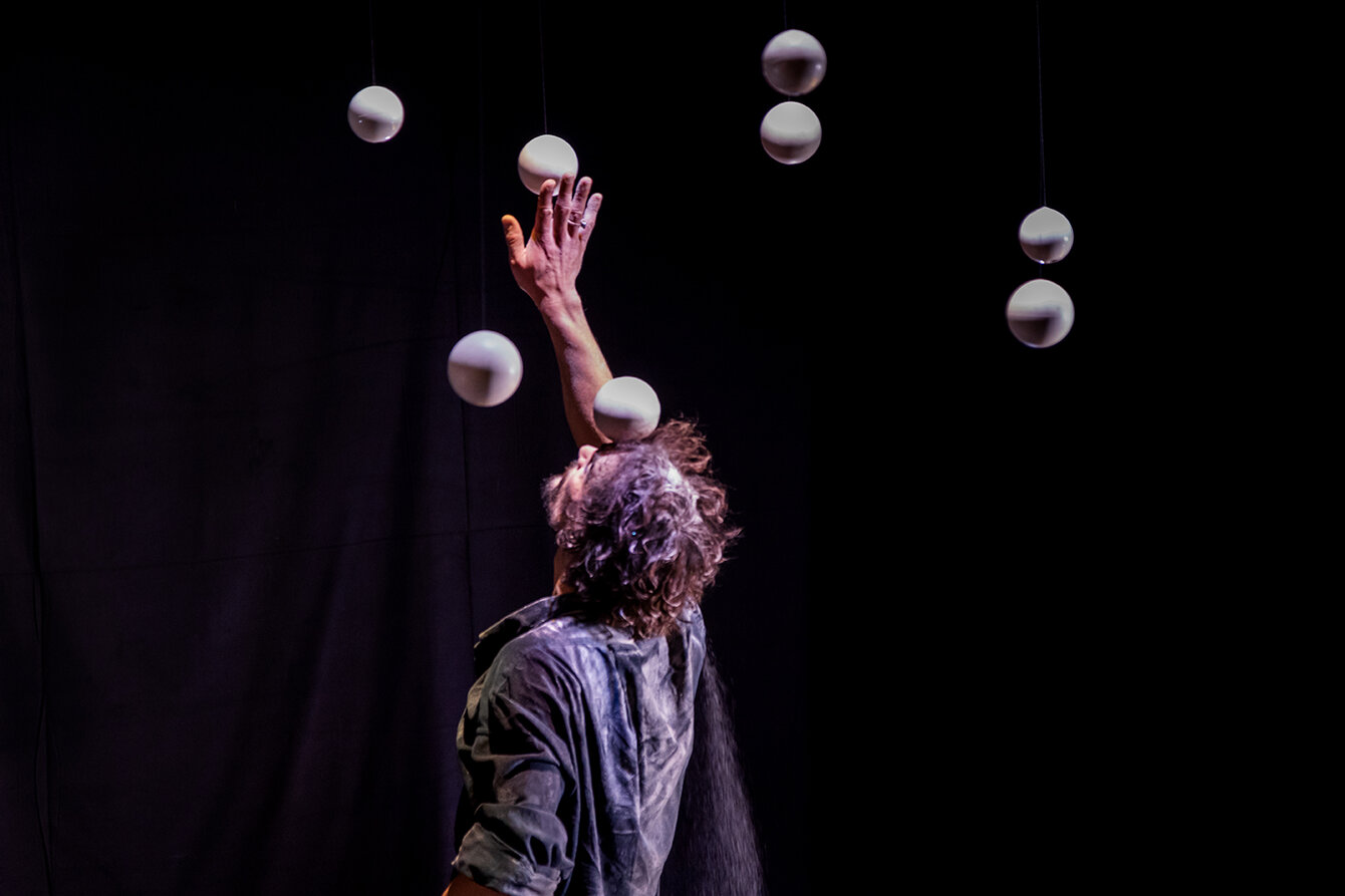 Juggler Tobias Piero Dohm performs in front of a dark background. He stretches his arm upwards. Several bright spherical objects float around him.