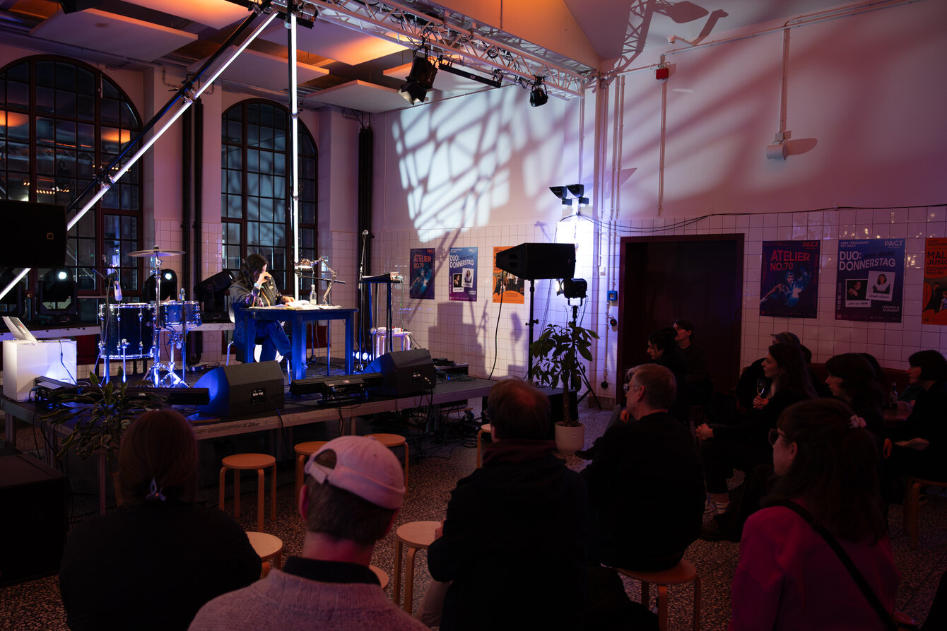 A woman is sitting on a stage in a large, open room. She is sitting at a table and reading from a book. The audience gathers around the stage and listens intently.