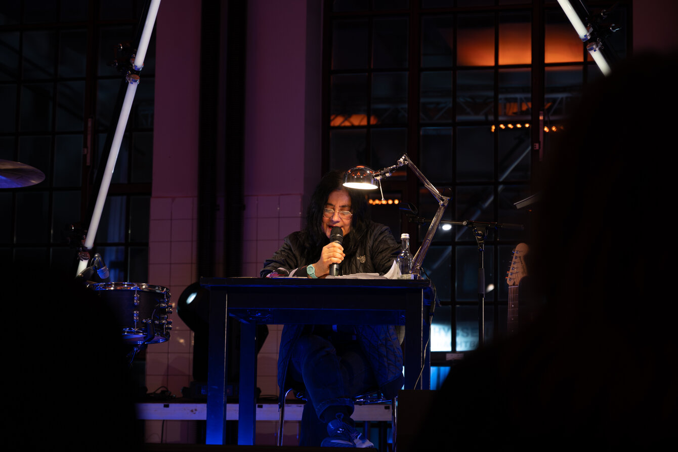 A woman is sitting at a table on a stage. A desk lamp illuminates the table and the woman's face. She is reading from a book.