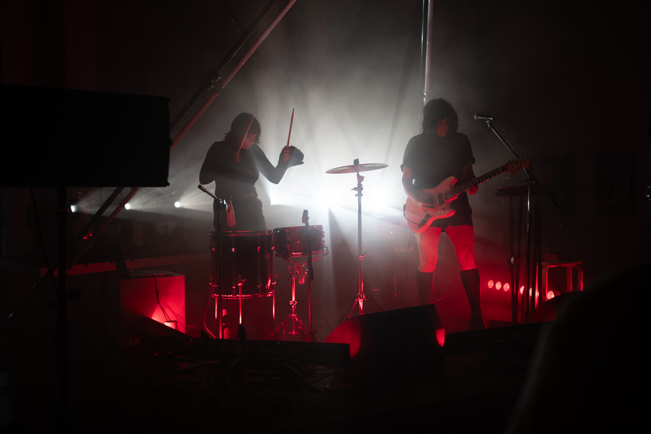 Two female musicians stand on a stage and are shrouded in red light. Their silhouettes are illuminated by a spotlight behind the stage.