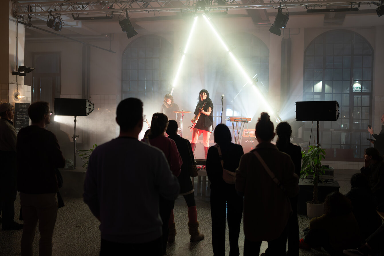 A band plays on a stage in a large, open room. The musicians are shrouded in fog and illuminated by fluorescent tubes behind the stage. The audience is spread out in front of the stage.