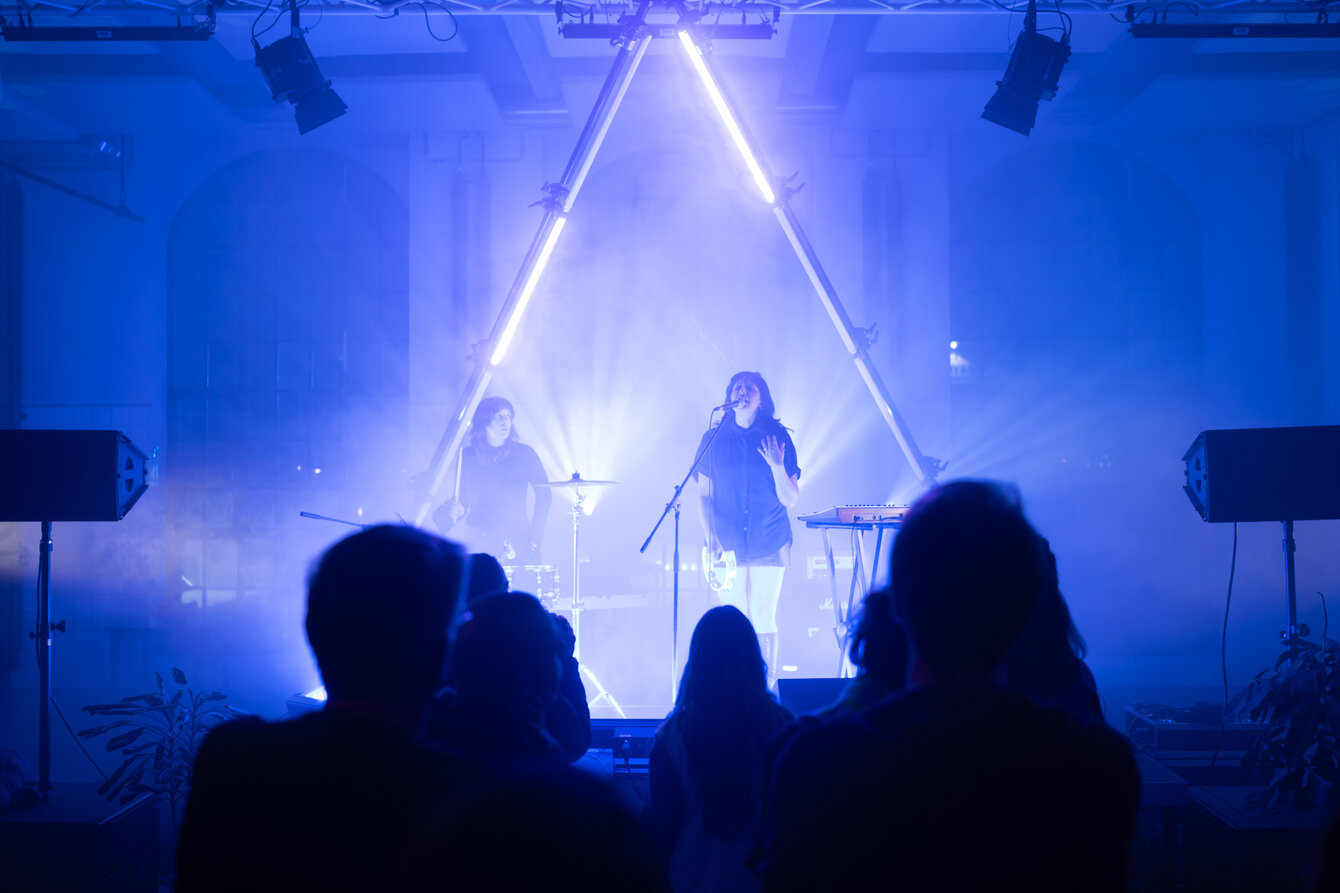 Two musicians stand on a stage. The room is shrouded in blue light and fog. Spotlights and fluorescent tubes cast dramatic light onto the stage.