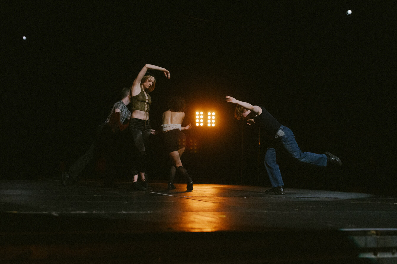 Several people dance on a dark stage. Three people are on the left, a hand gesture connects them to a single dancer on the right. An orange stage light shines softly through the center, the light is reflected on the dance floor.