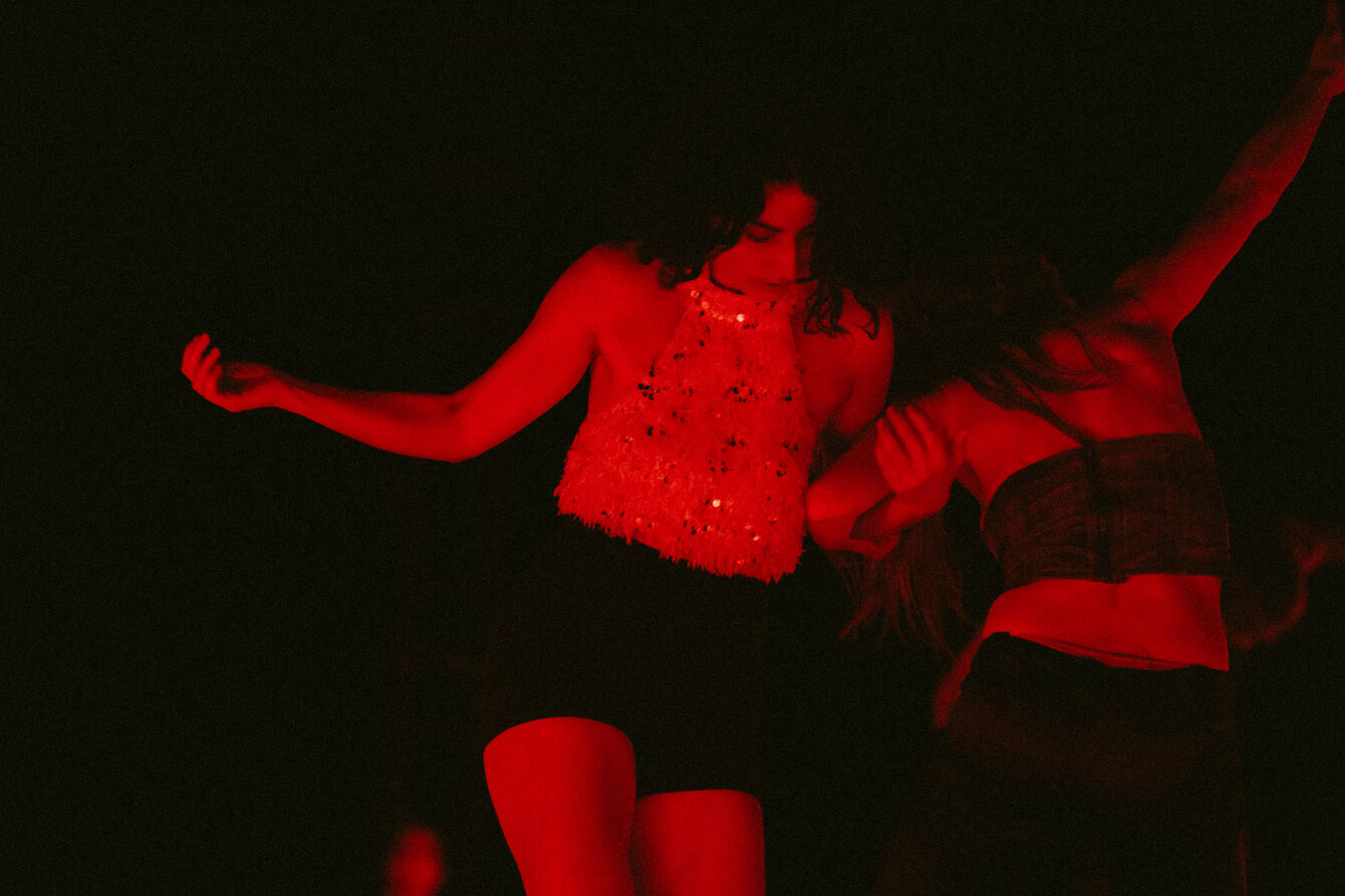 Two performers dance together in red light; they can be dimly recognized as they interlock their arms. The rest of the picture is dark.