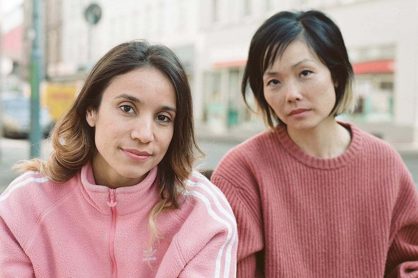 Ein Portrait von Bianca Pulungan (links) und Cinthia Nisiyama (rechts). Bianca hat schulterlange, braune Haare und trägt eine rosa Sportjacke. Cinthia hat kinnlange, dunkle Haare und trägt einen Pullover in der Farbe Altrosa. Sie befinden sich auf einer Straße. Beide blicken in die Kamera und lächeln leicht.